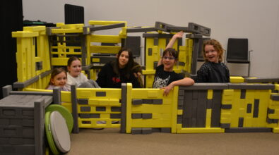 Grupa dzieci siedzi i bawi się wewnątrz dużej konstrukcji z klocków Mammutico w kolorach żółtym i szarym, przypominającej bazę kosmiczną z przejściami. Uczestnicy uśmiechają się, pozują do zdjęcia i unoszą ręce, wyraźnie dumni ze wspólnie stworzonej konstrukcji. W tle widoczna jest sala Mediateki z krzesłami, a cała scena oddaje radosną, integracyjną atmosferę kreatywnej zabawy i współpracy podczas zajęć.