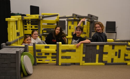 Grupa dzieci siedzi i bawi się wewnątrz dużej konstrukcji z klocków Mammutico w kolorach żółtym i szarym, przypominającej bazę kosmiczną z przejściami. Uczestnicy uśmiechają się, pozują do zdjęcia i unoszą ręce, wyraźnie dumni ze wspólnie stworzonej konstrukcji. W tle widoczna jest sala Mediateki z krzesłami, a cała scena oddaje radosną, integracyjną atmosferę kreatywnej zabawy i współpracy podczas zajęć.