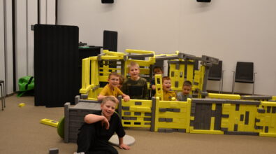 Grupa dzieci bawi się w sali Mediateki, pozując do zdjęcia przy dużej konstrukcji z klocków Mammutico w kolorach żółtym i szarym, przypominającej bazę kosmiczną. Uczestnicy z uśmiechami spoglądają w stronę aparatu, część z nich znajduje się wewnątrz budowli, a jedno z dzieci siedzi na podłodze przed konstrukcją. W tle widoczne są krzesła i elementy wyposażenia sali, a całość oddaje radosną atmosferę wspólnej zabawy i współpracy podczas zajęć feryjnych.