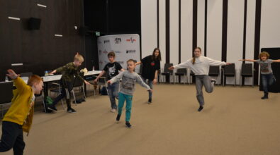 Grupa dzieci w różnym wieku bierze udział w zabawie ruchowej w sali Mediateki, wykonując ćwiczenia z rozłożonymi rękami i utrzymując równowagę podczas ruchu. Uczestnicy są rozstawieni w przestrzeni sali, a ich aktywność wskazuje na zajęcia animacyjne. W tle widoczne są stoły, krzesła oraz elementy wyposażenia sali konferencyjnej, a całość oddaje energiczną i radosną atmosferę wspólnej zabawy podczas ferii.