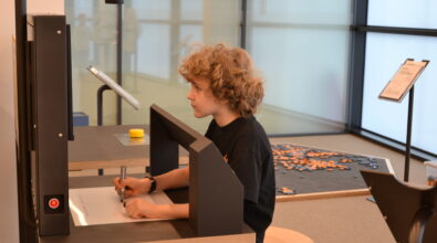 Chłopiec z kręconymi włosami siedzi przy stanowisku edukacyjnym w Mediatece i rysuje markerem na podświetlanej tablicy, skupiony na wykonywanym zadaniu. W tle widoczne są nowoczesne elementy przestrzeni edukacyjnej, w tym stanowiska do samodzielnej pracy i strefa z rozsypanymi klockami, co podkreśla twórczy i aktywny charakter zajęć prowadzonych podczas ferii zimowych.