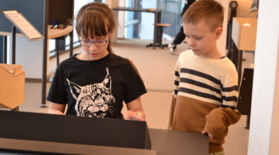 Dwoje dzieci stoi przy interaktywnym stanowisku edukacyjnym w Mediatece i wspólnie koncentruje się na wykonywanym zadaniu. Dziewczynka w okularach obsługuje panel, uważnie śledząc wyświetlane treści, a chłopiec stojący obok obserwuje jej działania z zainteresowaniem. W tle widoczna jest nowoczesna przestrzeń edukacyjna z kolejnymi stanowiskami do nauki przez działanie, sprzyjająca współpracy i odkrywaniu w trakcie zajęć.