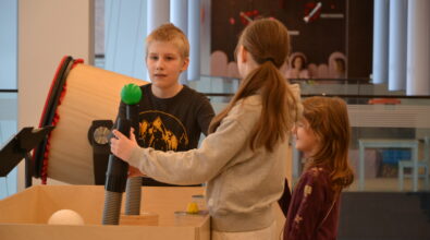 Troje dzieci uczestniczy w zajęciach w strefie SOWA w Mediatece, wspólnie korzystając z interaktywnego stanowiska edukacyjnego. Chłopiec trzyma element instalacji, a dziewczynka reguluje urządzenie z zieloną kulą, podczas gdy młodsza dziewczynka obserwuje doświadczenie z zainteresowaniem.