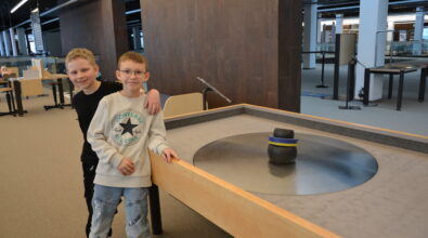 Dwóch chłopców stoi przy interaktywnym stanowisku edukacyjnym w Mediatece, przy którym znajduje się okrągły stół z obracającym się elementem demonstracyjnym. Jeden z chłopców obejmuje kolegę ramieniem, obaj uśmiechają się i spoglądają w stronę aparatu. W tle widoczna jest przestronna, nowoczesna sala strefy SOWA z kolejnymi stanowiskami do nauki przez działanie, sprzyjająca wspólnemu odkrywaniu i zabawie.