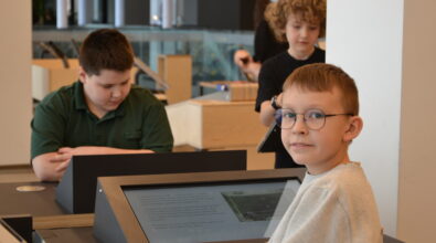 Troje dzieci uczestniczy w zajęciach edukacyjnych w nowoczesnej przestrzeni Mediateki, korzystając z interaktywnego stanowiska multimedialnego. Na pierwszym planie chłopiec w okularach stoi przy ekranie dotykowym i spogląda w stronę aparatu, a w tle pozostali uczestnicy skupiają się na pracy przy sąsiednich stanowiskach. Wnętrze jest jasne i przestronne, z widocznymi elementami strefy edukacyjnej sprzyjającej nauce przez działanie i wspólnemu odkrywaniu.