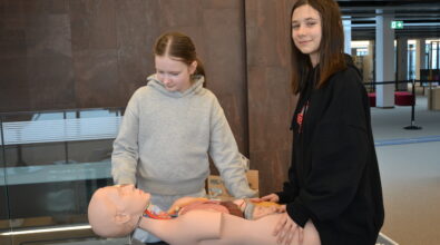 Dwie dziewczynki uczestniczą w zajęciach edukacyjnych w Mediatece, pracując przy realistycznym fantomie anatomicznym przedstawiającym ciało człowieka z widocznymi narządami wewnętrznymi. Jedna z uczestniczek uważnie obserwuje model, druga trzyma dłoń na tułowiu fantomu i spogląda w stronę aparatu.