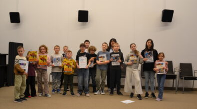 Grupa dzieci w różnym wieku stoi ustawiona w jednym rzędzie w sali Mediateki i pozuje do wspólnego zdjęcia, trzymając w dłoniach dyplomy oraz kolorowe książki. Uczestnicy uśmiechają się do aparatu, a za nimi widoczna jest jasna ściana sali oraz kilka krzeseł, co podkreśla oficjalny, ale radosny charakter podsumowania ferii.