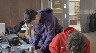 Troje młodych uczestników pracujących przy stole w przestrzeni bibliotecznej. Dwie osoby pochylają się nad elementami elektronicznymi i łączą podzespoły, a obok znajduje się otwarty laptop oraz pojemnik z drobnymi częściami. W tle widoczne są przeszklone drzwi z oznaczeniem pracowni multimedialnej oraz regały z książkami. Fotografia przedstawia etap konstruowania i testowania urządzenia wykorzystywanego w projekcie dotyczącym monitorowania jakości powietrza.