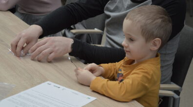 Mały chłopczyk siedzi przy stole obok dorosłego opiekuna i z zainteresowaniem trzyma w dłoni niewielką diodę podłączoną do przewodu. Dorosły pomaga mu przygotować elementy potrzebne do wykonania prostego doświadczenia, wyjmując części z niewielkiego opakowania. Na blacie leży karta z instrukcją, co wskazuje na uporządkowany i edukacyjny charakter zajęć.