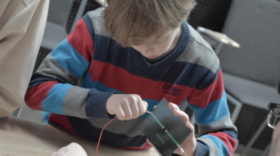Chłopiec siedzi przy stole i z dużym skupieniem podłącza przewody do niewielkiego panelu fotowoltaicznego. W dłoniach trzyma płytkę z oznaczeniami biegunów, do której za pomocą krokodylków mocuje czerwony i zielony przewód, tworząc prosty obwód elektryczny. Obok widoczna jest dłoń dorosłego, który wspiera dziecko w prawidłowym wykonaniu zadania.