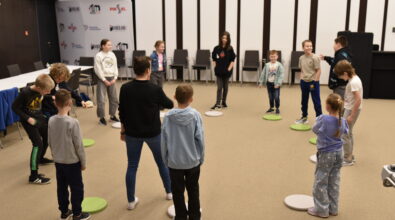 Dzieci stoją w kręgu w sali Mediateki i biorą udział w zabawie integracyjnej prowadzonej przez animatorkę. Uczestnicy wykonują polecenia i reagują na sygnały, stojąc na kolorowych podkładkach rozmieszczonych na podłodze. W tle widoczne są krzesła oraz ścianka z logotypami, a cała scena oddaje dynamiczną, wspólnotową atmosferę zajęć sprzyjających integracji i aktywności ruchowej.