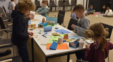 Dzieci stoją przy dużym stole w sali Mediateki i pracują nad zajęciami plastycznymi, wykorzystując papier kolorowy, karton, patyczki, kredki oraz drobne materiały dekoracyjne. Uczestnicy są skupieni na wycinaniu, klejeniu i konstruowaniu makiet, a na blacie widoczne są rozłożone przybory oraz rozpoczęte prace. W tle znajdują się krzesła i inni uczestnicy zajęć, co podkreśla warsztatowy, twórczy charakter wspólnej aktywności.