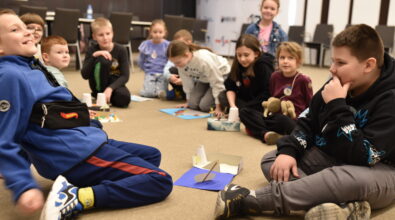 Grupa dzieci siedzi w kręgu na podłodze sali Mediateki i bierze udział w zajęciach integracyjno-edukacyjnych, rozmawiając i wspólnie wykonując zadania. Przed uczestnikami znajdują się proste prace plastyczne, papierowe elementy i drobne rekwizyty, a dzieci uważnie słuchają, obserwują i reagują na siebie nawzajem. Scena oddaje swobodną, przyjazną atmosferę zajęć, sprzyjającą współpracy, kreatywności i budowaniu relacji w grupie.