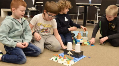 Czworo dzieci siedzi na podłodze sali Mediateki i wspólnie pracuje nad kreatywnymi makietami wykonanymi z papieru, kartonu i drobnych elementów plastycznych. Uczestnicy z zaangażowaniem układają i poprawiają szczegóły swoich prac, przedstawiających miniaturowe budowle i przestrzenie.