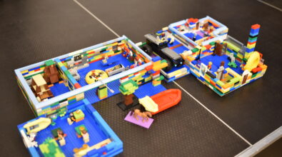 Na stole ustawiona jest rozbudowana makieta zbudowana z klocków LEGO, przedstawiająca połączone ze sobą pomieszczenia i przestrzenie z elementami wyposażenia, postaciami oraz pojazdami. Konstrukcja wykonana jest z kolorowych klocków, z wyraźnie zaznaczonymi ścianami, przejściami i detalami, takimi jak łódki, drzewa i figurki.