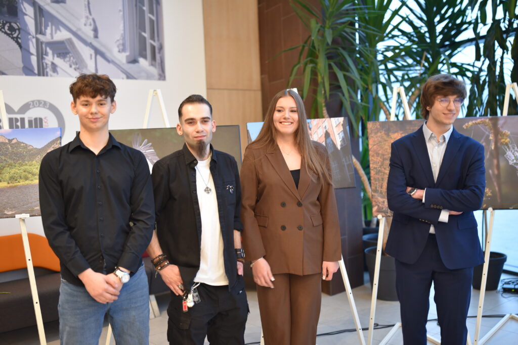 Czworo uczniów i absolwentów Zespołu Szkół Ponadpodstawowych nr 5 w Piotrkowie Trybunalskim „Fototechnika” pozuje do pamiątkowego zdjęcia podczas otwarcia wystawy w Mediatece 800-lecia. Młodzi twórcy stoją na tle swoich wielkoformatowych fotografii ustawionych na sztalugach, prezentujących m.in. pejzaże i architekturę. W tle widoczne jest nowoczesne wnętrze Mediateki z roślinnością i przeszkloną ścianą, podkreślające uroczysty charakter wydarzenia.