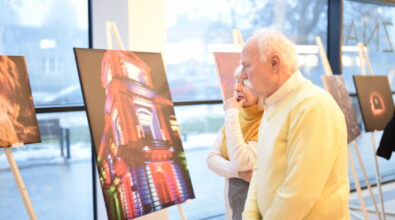 Para seniorów ogląda jedną z fotografii prezentowanych podczas otwarcia wystawy uczniów i absolwentów Zespołu Szkół Ponadpodstawowych nr 5 w Piotrkowie Trybunalskim „Fototechnika” w Mediatece 800-lecia. Na pierwszym planie widoczna jest wielkoformatowa fotografia przedstawiająca iluminowaną architekturę w intensywnych barwach, ustawiona na sztaludze przy przeszklonej ścianie budynku. Kobieta i mężczyzna stoją blisko ekspozycji, w skupieniu analizując detale pracy. W tle widać kolejne fotografie rozmieszczone wzdłuż okien oraz jasne, nowoczesne wnętrze Mediateki.