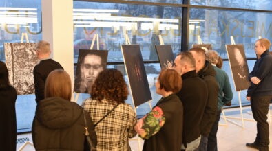 Goście zgromadzeni w Mediatece 800-lecia oglądają czarno-białe fotografie prezentowane podczas otwarcia wystawy „Fototechnika”. Prace ustawione na białych sztalugach tworzą ekspozycję wzdłuż przeszklonej ściany budynku z widocznym napisem Mediateki. Uczestnicy wydarzenia stoją w skupieniu przed portretami i artystycznymi ujęciami, wymieniając się wrażeniami. Za oknami widoczny jest zimowy krajobraz, który stanowi naturalne tło dla prezentowanych prac.