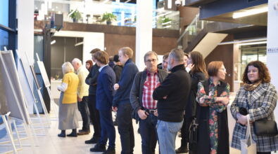 Grupa gości ogląda fotografie prezentowane podczas otwarcia wystawy uczniów i absolwentów Zespołu Szkół Ponadpodstawowych nr 5 w Piotrkowie Trybunalskim „Fototechnika” w Mediatece 800-lecia. Prace ustawione na białych sztalugach tworzą ekspozycję wzdłuż przestronnego holu. Uczestnicy wydarzenia stoją w skupieniu przed zdjęciami, prowadzą rozmowy i wymieniają się wrażeniami. W tle widoczne jest nowoczesne wnętrze Mediateki z antresolą, przeszklonymi ścianami i liniowym oświetleniem sufitowym.