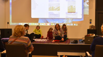 Na zdjęciu uczestnicy spotkania Klubu Podróżnika siedzą na scenie na kolorowym dywanie, słuchając opowieści prowadzącej. W wydarzeniu biorą udział dzieci oraz dorośli, którzy wspólnie uczestniczą w prezentacji. W tle widoczny jest duży ekran z wyświetlaną prezentacją multimedialną przedstawiającą zdjęcia z podróży. Przed sceną siedzą słuchacze, obserwując przebieg spotkania.