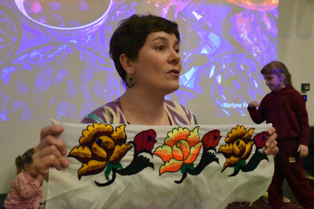 Na zdjęciu podróżniczka Martyna Kwiatkowska prezentuje uczestnikom spotkania ozdobną tkaninę z kolorowym, kwiatowym haftem. Prelegentka trzyma materiał w dłoniach i opowiada o jego pochodzeniu oraz znaczeniu w kulturze odwiedzanego kraju. W tle widoczny jest ekran z wyświetlaną prezentacją multimedialną ze zdjęciami z podróży, a obok sceny znajdują się dzieci uczestniczące w wydarzeniu.