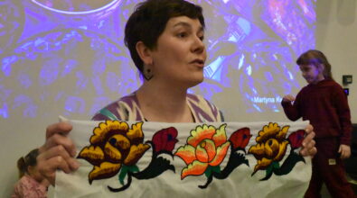 Na zdjęciu podróżniczka Martyna Kwiatkowska prezentuje uczestnikom spotkania ozdobną tkaninę z kolorowym, kwiatowym haftem. Prelegentka trzyma materiał w dłoniach i opowiada o jego pochodzeniu oraz znaczeniu w kulturze odwiedzanego kraju. W tle widoczny jest ekran z wyświetlaną prezentacją multimedialną ze zdjęciami z podróży, a obok sceny znajdują się dzieci uczestniczące w wydarzeniu.