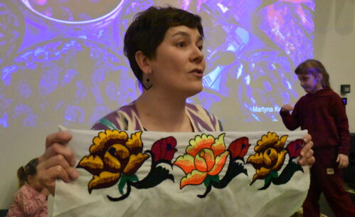 Na zdjęciu podróżniczka Martyna Kwiatkowska prezentuje uczestnikom spotkania ozdobną tkaninę z kolorowym, kwiatowym haftem. Prelegentka trzyma materiał w dłoniach i opowiada o jego pochodzeniu oraz znaczeniu w kulturze odwiedzanego kraju. W tle widoczny jest ekran z wyświetlaną prezentacją multimedialną ze zdjęciami z podróży, a obok sceny znajdują się dzieci uczestniczące w wydarzeniu.