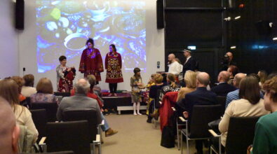 Na zdjęciu widoczna jest scena podczas spotkania Klubu Podróżnika w Mediatece 800-lecia. Na podwyższeniu stoją prowadząca oraz uczestnicy prezentacji ubrani w tradycyjne stroje inspirowane kulturą Tadżykistanu. W wydarzeniu aktywnie uczestniczą także dzieci zaproszone na scenę. W tle wyświetlana jest prezentacja multimedialna przedstawiająca zdjęcie tradycyjnych potraw, stanowiących element kultury i codziennego życia mieszkańców regionu. Publiczność siedząca na widowni z zainteresowaniem obserwuje pokaz.