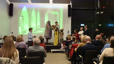 Na zdjęciu uczestnicy spotkania Klubu Podróżnika siedzą na widowni i obserwują prezentację odbywającą się na scenie. Na podwyższeniu stoją dzieci oraz prowadząca, która prezentuje elementy tradycyjnego stroju i tkanin związanych z kulturą Tadżykistanu. W tle wyświetlana jest prezentacja multimedialna przedstawiająca fotografie z podróży. Publiczność z zainteresowaniem śledzi pokaz, a scena ma charakter interaktywny – część uczestników została zaproszona do wspólnej prezentacji.