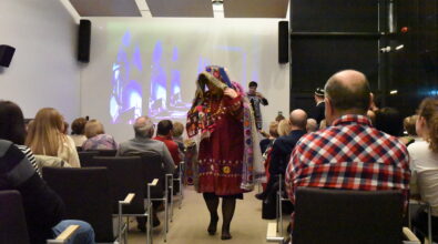 Na zdjęciu uczestniczka spotkania Klubu Podróżnika przechodzi między rzędami widowni ubrana w tradycyjny, bogato zdobiony strój inspirowany kulturą Tadżykistanu. Na scenie w tle widoczna jest prowadząca prezentację, a za nią ekran z wyświetlanymi zdjęciami z podróży. Publiczność siedząca na sali obserwuje pokaz, który stanowi część interaktywnej prezentacji podczas spotkania podróżniczego.