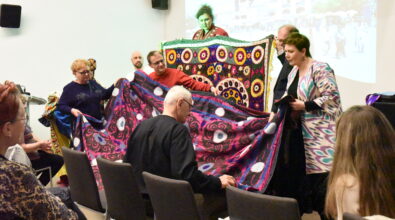 Na zdjęciu uczestnicy spotkania Klubu Podróżnika wspólnie prezentują duże, kolorowe tkaniny ozdobione bogatymi wzorami i haftami charakterystycznymi dla kultury Azji Centralnej. Kilka osób rozkłada materiał, aby zaprezentować jego wielkość oraz detale zdobień zgromadzonej publiczności. W tle widoczny jest ekran z wyświetlaną prezentacją multimedialną z podróży, a na widowni siedzą uczestnicy spotkania obserwujący pokaz.