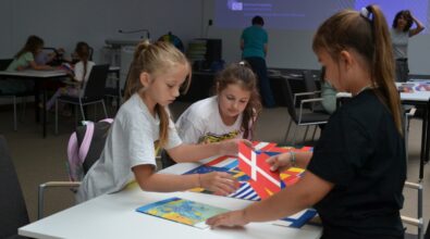 Trzy dziewczynki biorą udział w warsztatach edukacyjnych, układając puzzle z flagami państw europejskich. Pracują przy jednym ze stołów w przestronnej sali, w tle której widoczna jest prezentacja dotycząca Unii Europejskiej. Dzieci wykazują zaangażowanie i skupienie, co świadczy o dużym zainteresowaniu tematem. Zajęcia miały na celu przybliżenie młodym uczestnikom wiedzy o krajach Europy oraz promowanie wartości europejskich w przystępny i atrakcyjny sposób. To jedno z wielu wartościowych wydarzeń zorganizowanych w ramach wakacji z Mediateką.