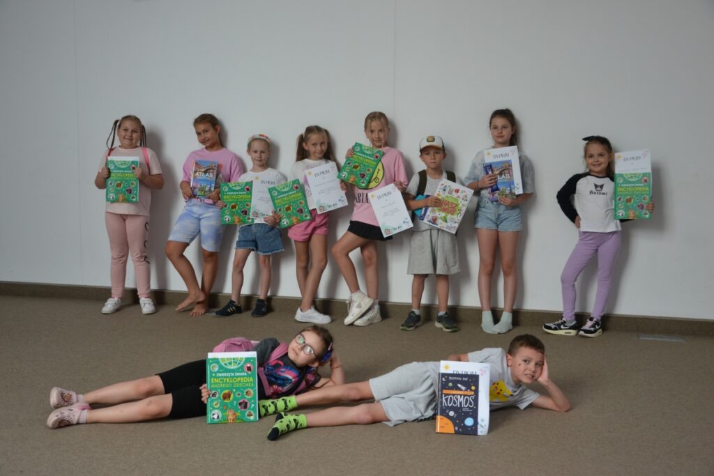 Grupa dzieci dumnie prezentujących dyplomy i książkowe upominki – nagrody za udział w wakacyjnych zajęciach organizowanych przez Mediatekę 800-lecia. Dziesięcioro dzieci stoi na tle jasnej ściany, a dwoje leży na podłodze z uśmiechem, eksponując swoje nagrody. W rękach uczestników znajdują się kolorowe encyklopedie, atlasy i książki edukacyjne, co świadczy o rozwijającym i wartościowym charakterze zajęć. Zdjęcie emanuje radością i satysfakcją z dobrze spędzonego czasu.