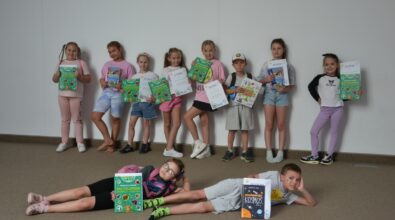 Grupa dzieci dumnie prezentujących dyplomy i książkowe upominki – nagrody za udział w wakacyjnych zajęciach organizowanych przez Mediatekę 800-lecia. Dziesięcioro dzieci stoi na tle jasnej ściany, a dwoje leży na podłodze z uśmiechem, eksponując swoje nagrody. W rękach uczestników znajdują się kolorowe encyklopedie, atlasy i książki edukacyjne, co świadczy o rozwijającym i wartościowym charakterze zajęć. Zdjęcie emanuje radością i satysfakcją z dobrze spędzonego czasu.