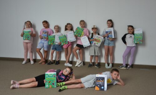 Grupa dzieci dumnie prezentujących dyplomy i książkowe upominki – nagrody za udział w wakacyjnych zajęciach organizowanych przez Mediatekę 800-lecia. Dziesięcioro dzieci stoi na tle jasnej ściany, a dwoje leży na podłodze z uśmiechem, eksponując swoje nagrody. W rękach uczestników znajdują się kolorowe encyklopedie, atlasy i książki edukacyjne, co świadczy o rozwijającym i wartościowym charakterze zajęć. Zdjęcie emanuje radością i satysfakcją z dobrze spędzonego czasu.