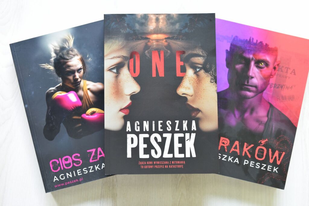 Trzy egzemplarze książki autorstwa Agnieszka Peszek ustawione obok siebie, lekko pod kątem. W centrum znajduje się książka z wyraźnym tytułem „ONE”. Okładka przedstawia zbliżenia twarzy młodych osób skierowanych w różnych kierunkach, utrzymane w chłodnej kolorystyce. Po lewej stronie widoczna jest książka w odcieniach różu i fioletu z fragmentem tytułu „…es za”, a po prawej egzemplarz w ciemniejszych, purpurowych barwach z widocznym fragmentem tytułu „Rak…”. Wszystkie okładki tworzą spójną serię graficzną z portretami bohaterów i intensywnymi kolorami tła.