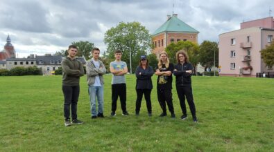 Sześcioosobowa grupa stojąca na zielonej trawie. Uczestnicy ustawieni są w jednym rzędzie, z rękami skrzyżowanymi na piersiach, i patrzą w stronę aparatu. W tle znajduje się zabytkowy budynek z cegły z zielonym dachem oraz drzewa o bujnych koronach. Po prawej stronie widoczny jest jasnoróżowy budynek mieszkalny z balkonami. Nad całą sceną rozciąga się pochmurne, zachmurzone niebo, które nadaje fotografii nieco surowy, jesienny charakter.