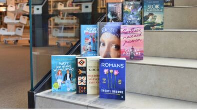 Na zdjęciu widzimy stos książek ułożonych na schodach w bibliotece. W tle znajduje się przestrzeń biblioteczna z regałami pełnymi książek oraz stanowiskami komputerowymi. Na pierwszym planie widoczne są okładki różnych powieści, m.in.: „Patrz ze mną w gwiazdy” Anny Szafrańskiej, „Romans” Sheryl Browne, „Pięcioro małych Indian” Michelle Good, „Spotkasz mnie nad jeziorem” Carley Fortune, „Oczy Mony” i inne. Na dole zdjęcia umieszczono logotypy: Narodowego Programu Rozwoju Czytelnictwa 2.0, Ministerstwa Kultury i Dziedzictwa Narodowego oraz Biblioteki Narodowej. Widnieje też informacja o dofinansowaniu ze środków Ministra Kultury w ramach realizacji NPRCz 2.0 na lata 2021–2025.