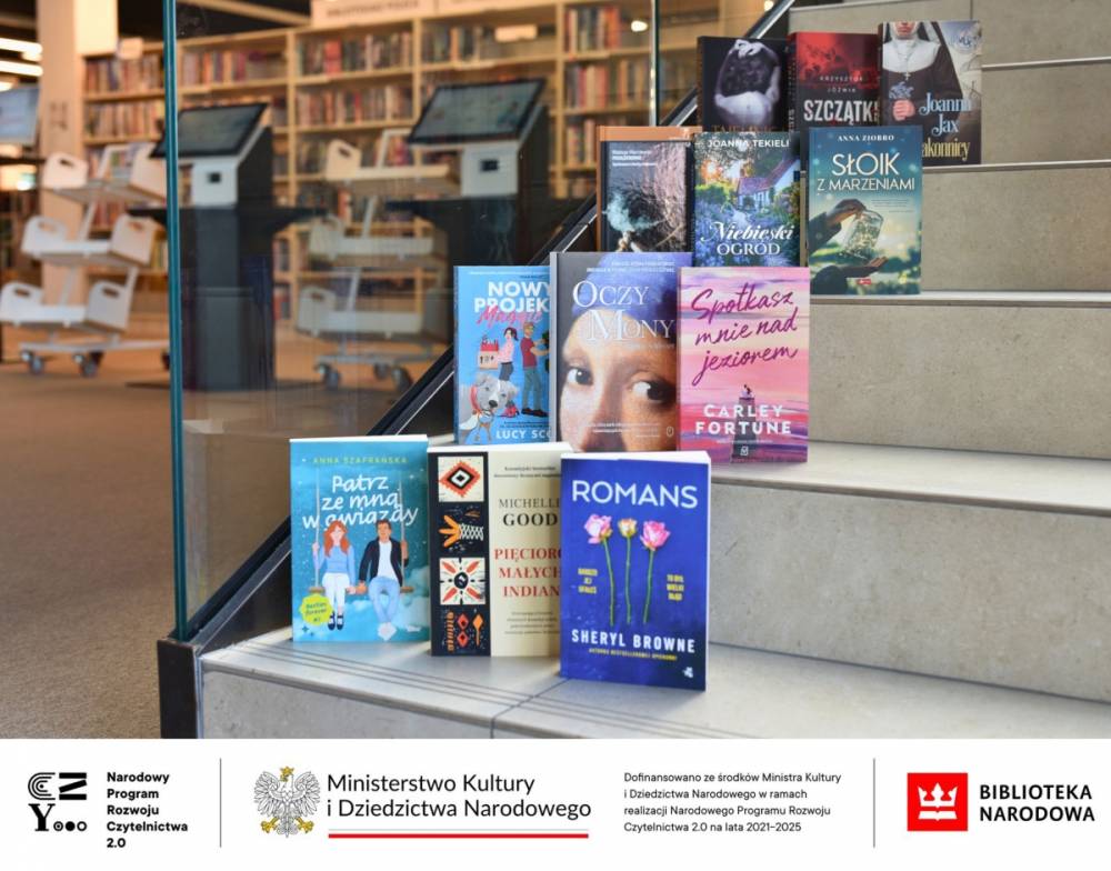 Na zdjęciu widzimy stos książek ułożonych na schodach w bibliotece. W tle znajduje się przestrzeń biblioteczna z regałami pełnymi książek oraz stanowiskami komputerowymi. Na pierwszym planie widoczne są okładki różnych powieści, m.in.: „Patrz ze mną w gwiazdy” Anny Szafrańskiej, „Romans” Sheryl Browne, „Pięcioro małych Indian” Michelle Good, „Spotkasz mnie nad jeziorem” Carley Fortune, „Oczy Mony” i inne. Na dole zdjęcia umieszczono logotypy: Narodowego Programu Rozwoju Czytelnictwa 2.0, Ministerstwa Kultury i Dziedzictwa Narodowego oraz Biblioteki Narodowej. Widnieje też informacja o dofinansowaniu ze środków Ministra Kultury w ramach realizacji NPRCz 2.0 na lata 2021–2025.