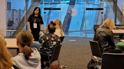 Nowoczesna sala ze szklanymi ścianami, w której przy stołach siedzi grupa ludzi. W tle widać ekran projektowy i osobę prowadzącą zajęcia.