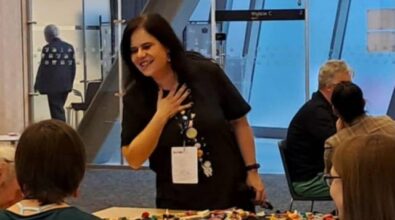 Na zdjęciu widnieją osoby siedzące wokół stołu, który jest pokryty różnokolorowymi elementami LEGO. Jedna osoba stoi i prowadzić prezentację. Tło obejmuje szyby szklane i drzwi, co wskazuje na nowoczesną, otwartą przestrzeń. Widać również ekran projekcyjny.