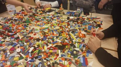 Zbliżenie na stół na którym rozłożono bardzo dużą ilość klocków LEGO. Przy stole widać siedzące osoby, które próbują coś zbudować.