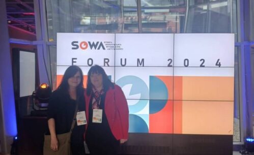 dwie osoby stojące przed dużym ekranem wyświetlającym informacje o "SOWA FORUM 2024". Logo i abstrakcyjne wzory geometryczne dodatkowo zdobią ekran. Wydarzenie odbywa się w lokalu o nowoczesnym, przemysłowym stylu. Obie kobiety noszą opaski z nazwiskami, co wskazuje, że są uczestnikami forum.