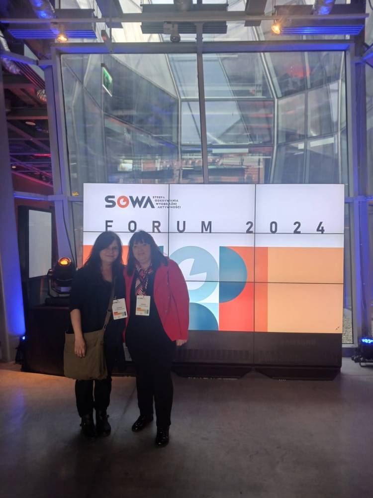 dwie osoby stojące przed dużym ekranem wyświetlającym informacje o "SOWA FORUM 2024". Logo i abstrakcyjne wzory geometryczne dodatkowo zdobią ekran. Wydarzenie odbywa się w lokalu o nowoczesnym, przemysłowym stylu. Obie kobiety noszą opaski z nazwiskami, co wskazuje, że są uczestnikami forum.