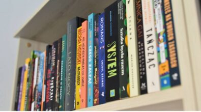 Na zdjęciu widzimy fragment regału z książkami w bibliotece. Półka jest wypełniona kolorowymi grzbietami książek. Wśród widocznych tytułów znajdują się m.in. „System” Sheryla Browne, „Nie wszystko muszą cię lubić” Michała Fegurskiego, „Od czego zacząć” oraz „Tajemnica Gai”. Na dole zdjęcia umieszczono logotypy: Narodowego Programu Rozwoju Czytelnictwa 2.0, Ministerstwa Kultury i Dziedzictwa Narodowego oraz Biblioteki Narodowej. Widnieje również informacja o dofinansowaniu w ramach NPRCz 2.0 na lata 2021–2025. Kadr ukazuje uporządkowane i estetyczne ułożenie książek w jasnym, nowoczesnym wnętrzu.