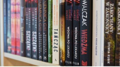 Na zdjęciu znajduje się półka z książkami w bibliotece, wypełniona różnorodnymi tytułami. Widoczne książki to m.in. „Tajemnica Gai”, „Wdowa po lekarzu” Daniela Hursta, „Wiedźma” Marcina Walczaka oraz „Zakochany w zakonnicy” Katarzyny Kazimierowskiej. Na dole zdjęcia umieszczono logotypy Narodowego Programu Rozwoju Czytelnictwa 2.0, Ministerstwa Kultury i Dziedzictwa Narodowego oraz Biblioteki Narodowej. Informacja podkreśla, że dofinansowanie pochodzi z programu NPRCz 2.0 na lata 2021–2025. Kadr przedstawia uporządkowaną biblioteczną półkę w jasnym wnętrzu.