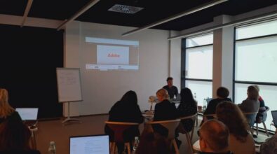 Sala lekcyjna z prezentacją. Osoba siedzi przed ekranem projektora wyświetlającym logo Adobe. Kilka osób siedzi przy stołach z laptopami. Po lewej stronie widoczna jest tablica.