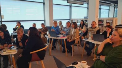 Grupa osób siedzi w dobrze oświetlonym pomieszczeniu podczas spotkania lub warsztatu. Wszyscy mają laptopy i notatniki na stołach przed sobą i wydają się zaangażowani i uważni. Duże okna w tle wpuszczają naturalne światło.