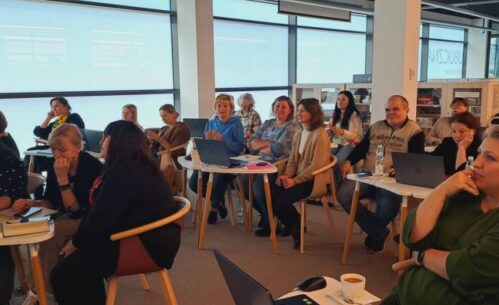 Grupa osób siedzi w dobrze oświetlonym pomieszczeniu podczas spotkania lub warsztatu. Wszyscy mają laptopy i notatniki na stołach przed sobą i wydają się zaangażowani i uważni. Duże okna w tle wpuszczają naturalne światło.