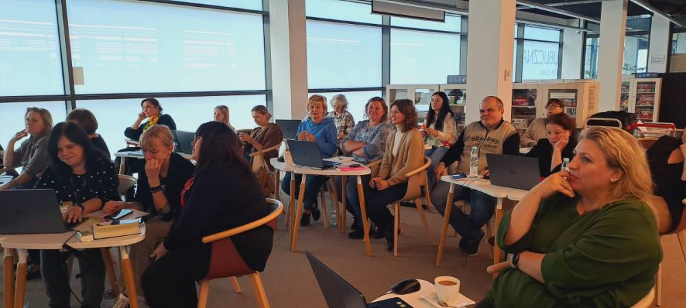 Grupa osób siedzi w dobrze oświetlonym pomieszczeniu podczas spotkania lub warsztatu. Wszyscy mają laptopy i notatniki na stołach przed sobą i wydają się zaangażowani i uważni. Duże okna w tle wpuszczają naturalne światło.